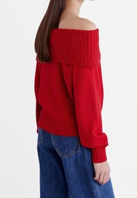 Femme aux longs cheveux bruns portant un pull en maille rouge à épaules dénudées et un jean en denim bleu, debout devant un fond blanc.