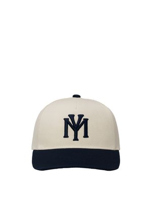 Kvinde iført en beige og marineblå kasket, gyldne hoops-øreringe, en grå sweatshirt med rødt og blåt stjernedesign og et bundet marineblåt mønstret halstørklæde.