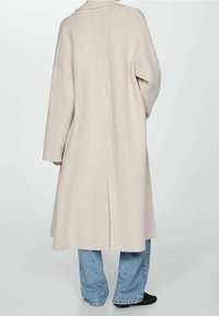 Pitkä beige takki, valmistettu pehmeästä materiaalista, rento istuvuus, leveät hihat ja keskiselän keskisauma, yhdistetty löysiin sinisiin farkkuihin.