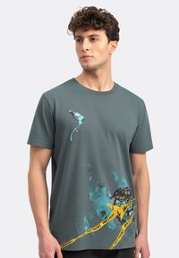 Grijze katoenen T-shirt met een ronde hals, met een grafische afbeelding van een verfpot en spetters in blauw, geaccentueerd met een gele tape-ontwerp "WET PAINT".