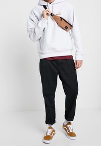 Sudadera gris clara con un bolsillo canguro, pantalones oscuros ajustados, zapatos beige y una riñonera de pana marrón con cremallera.