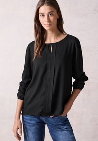 Blusa nera a maniche lunghe con scollatura rotonda, caratterizzata da una dettaglio a chiave frontale e un leggero motivo testurizzato. Abbinata a jeans blu.