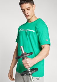 Vihreä puuvillainen t-paita, jossa on valkoinen "Champion" -logo edessä. Kantaja pitää pitkää tankoa, jossa on mustia painoja. Harmaat verkkarit näkyvät.