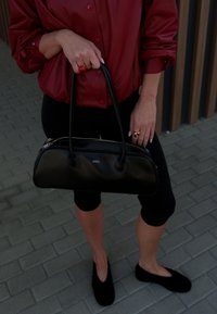 Czarna skórzana torba w zaokrąglonym kształcie, z podwójnymi uchwytami u góry, centralnym zapięciem na zamek, matowym wykończeniem, logo z przodu. Noszona z czerwoną bluzką i czarnymi legginsami.