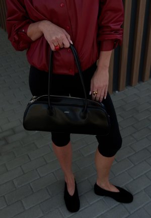 Czarna skórzana torba w zaokrąglonym kształcie, z podwójnymi uchwytami u góry, centralnym zapięciem na zamek, matowym wykończeniem, logo z przodu. Noszona z czerwoną bluzką i czarnymi legginsami.