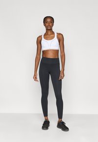 Vit sport-bh med enkel design, kombinerad med svart, midjehöga leggings. Modellen bär svarta träningsskor. Slät textur.