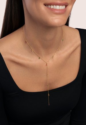 Goldene Halskette mit bunten Edelsteinakzenten, im Y-förmigen Design mit kleinen hängenden Elementen. Tragen mit einem schwarzen Oberteil.