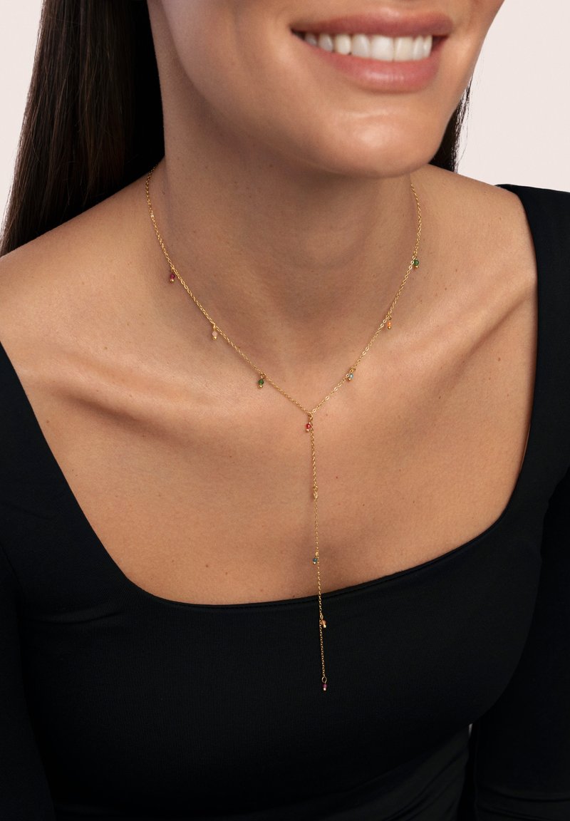 Collana in oro con accenti di pietre preziose colorate, design a forma di Y e piccoli elementi pendenti. Indossata con un top nero.