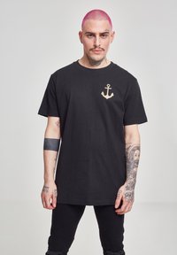 Černé bavlněné tričko s grafickým motivem zlatého kotvy na levé části hrudi. Model má tetování a nosí černé kalhoty.