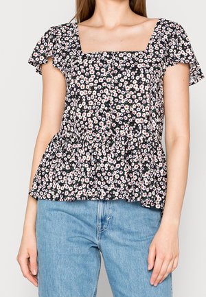 Kobieta w czarnej, białej i czerwonej kwiecistej bluzce z krótkim rękawem, z kwadratowym dekoltem oraz niebieskich dżinsach z wysokim stanem.