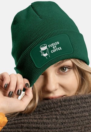 Green knitted beanie with ribbed texture and a patch reading "FUELED BY COFFEE." Close-up view of a person's hand and face partially concealed.