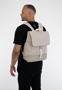 Világos bézs hátizsák hajtókával, állítható pántokkal és elülső csattal. Simasága, a talp körüli finom díszítés és minimalista dizájn jellemzi.