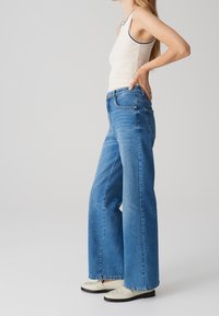 Vaqueros azules de pierna ancha hechos de mezclilla, con cintura alta y bolsillos laterales. Combinados con una camiseta sin mangas clara y zapatos blancos sin cordones.