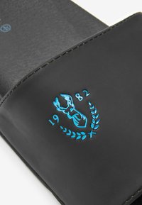Svart slidesandal med en texturerad yta. Har en blå broderad hummer och året "1982" omgivet av lagerblad.