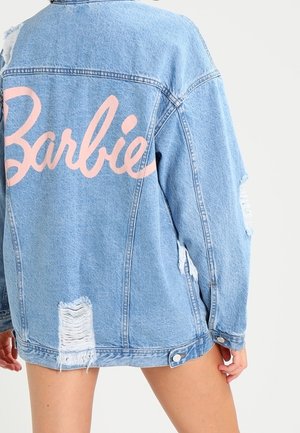 Jasnoniebieska znoszona kurtka dżinsowa z dużym różowym napisem „Barbie” na plecach, noszona przez osobę stojącą na jednolitym tle.
