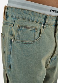 Hele sinised teksapüksid lähivõttega, kus on näha tagatasku, õmbluse detailid ja valge vöökoht "PEGADOR" logoga.