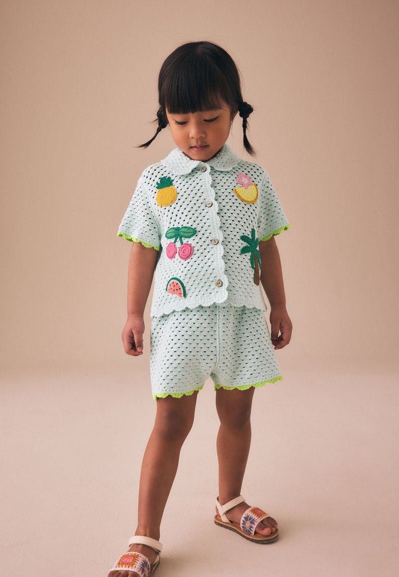 Niña joven con coletas lleva una camisa blanca de ganchillo y pantalones cortos con parches de frutas coloridas y palmeras y sandalias bordadas.