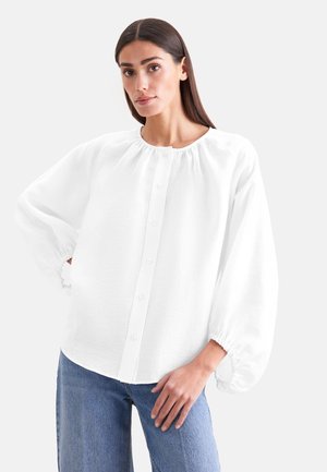 Vrouw met steil donker haar, gekleed in een losse witte blouse met knopen en geplooide manchetten en blauwe spijkerbroek, staand tegen een witte achtergrond.