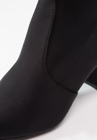 Bottine noire à la cheville avec bout pointu, tissu lisse et talon bloc, montrée en gros plan sur fond blanc.