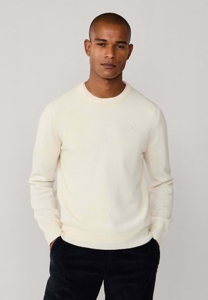 Cremfarbener gestrickter Pullover mit Rundhalsausschnitt und gerippten Bündchen. Verfügt über ein dezentes Logo auf der Brust. Kombiniert mit dunkler Kleidung.