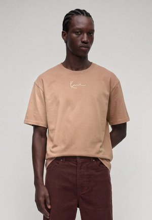 Jeune homme aux cheveux tressés portant une chemise beige à manches courtes avec des inscriptions blanches et un pantalon en velours côtelé marron foncé sur fond uni.