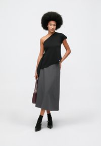 Top preto assimétrico com uma manga curta à esquerda, combinado com uma saia longa cinza. Modelo segurando uma bolsa bordô; sapatos pretos com detalhes.