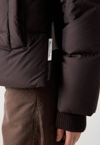 Veste rembourrée marron foncé avec fermeture éclair, dotée d'une étiquette de marque, de poignets côtelés et d'un tissu à texture lisse.