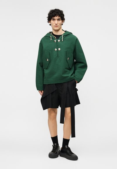 Νέος άνδρας που φοράει πλεκτό πράσινο φούτερ με κουκούλα και μεγάλα κουμπιά, μαύρο ασύμμετρο σορτς με μακριές λωρίδες υφάσματος, μαύρες κάλτσες και παπούτσια.