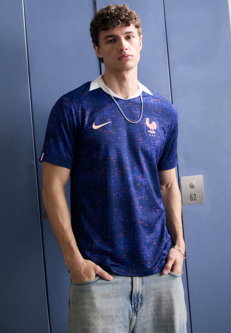 Blauwe Nike-jersey met rood en paars pixelpatroon, witte kraag en geborduurd hanenlogo. Draag met lichtblauwe jeans.