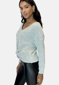 Lichtgrijze gebreide trui met een V-hals en een gedraaide voorkant. Gestructureerde stof en lange mouwen, gecombineerd met zwarte leggings.