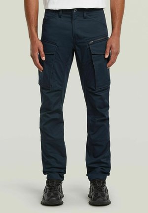 Uomo che indossa pantaloni cargo blu scuro con multiple tasche e sneakers nere, in piedi su uno sfondo semplice.