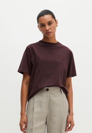 Femme aux cheveux foncés attachés en arrière portant un t-shirt à manches courtes violet foncé avec un détail en forme de cœur et un pantalon taille haute gris clair.