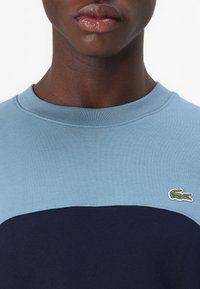 Felpa crewneck blu chiaro e navy, con tessuto texture e un piccolo logo di coccodrillo ricamato sul lato sinistro del petto.