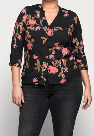 Frau trägt schwarze Bluse mit Knöpfen, verziert mit rosa-braunem Blumenmuster, und schwarze Jeans, steht vor schlichtem Hintergrund.