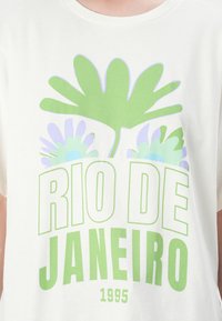 T-shirt en coton blanc ample avec un design imprimé vert et lavande, comportant des feuilles tropicales et la mention "RIO DE JANEIRO 1995" en lettres majuscules.