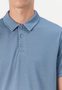 Teksturirana kratka polo majica s kratkimi rokavi v svetlo modri barvi, z zloženim ovratnikom, trojnim gumbom in vodoravno rebrasto vzorcem.
