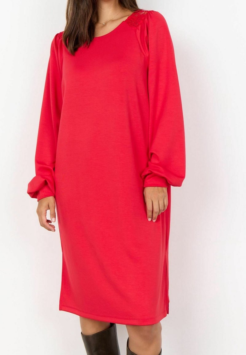 Femme portant une robe rouge à manches longues arrivant aux genoux avec des manches légèrement bouffantes et des bottes noires mi-mollet sur fond blanc.