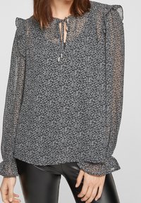 Blouse noire avec un délicat motif floral blanc, tissu transparent, bords de manches volants et un détail de col en lien avec des accents métalliques.