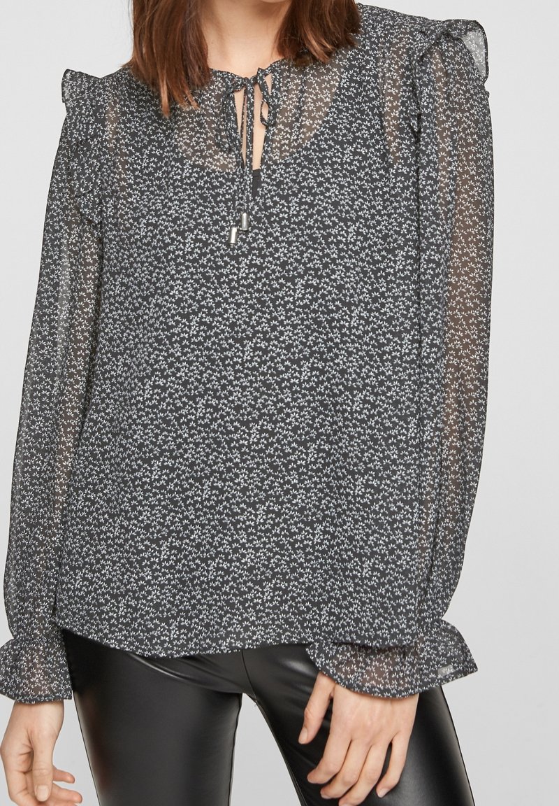 Blouse noire avec un délicat motif floral blanc, tissu transparent, bords de manches volants et un détail de col en lien avec des accents métalliques.
