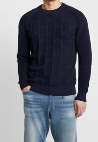 Marinblå tröja med ett strukturerat mönster av diamantformer, ribbade ärmar och halsringning samt en figurnära midja. Matchad med ljusblå jeans.