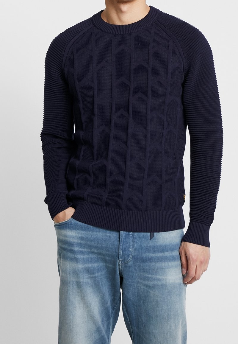 Marinblå tröja med ett strukturerat mönster av diamantformer, ribbade ärmar och halsringning samt en figurnära midja. Matchad med ljusblå jeans.