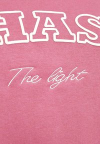 Roze stof met grote, witte blokletters en cursieve tekst. De tekst leest "CHAS" en "The light" in contrasterende borduurwerk. Zachte textuur.