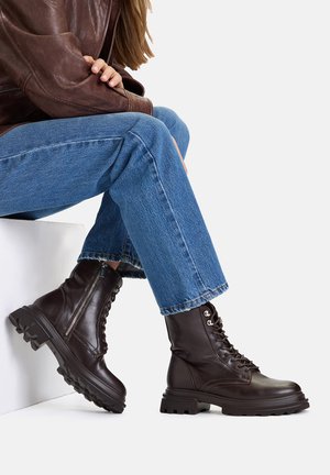 Brązowe skórzane buty bojowe z wiązaniem na sznurowadła i masywną podeszwą, wyposażone w boczny zamek, noszone z niebieskimi dżinsami. Gładka faktura i solidna konstrukcja.