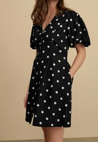 Abito nero con pois bianchi, dotato di maniche a sbuffo, scollatura a V e due tasche laterali. Tessuto liscio e apertura frontale con bottoni.