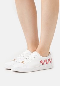 Baskets blanches en cuir avec inscription "KORS" rouge et accents à motifs damiers, dotées d'un laçage plat et de détails subtils en or.