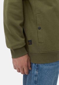 Sweat-shirt vert olive avec des poignets côtelés, une poche latérale et un détail de bouton pression noir ; associé à un jean en denim bleu clair, à coupe décontractée.