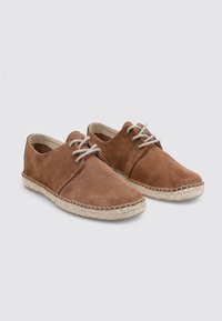Zapatos de gamuza marrón con un perfil bajo, punta redonda y suela de yute estilo espadrilles en color beige. Presentan cordones grises y detalles de costura mínimos.