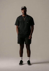 Black t-shirt and cargo shorts, both in a plain design; black cap; gold jewelry; green shoes; white socks with a logo. Neutral background.