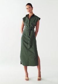 Abito verde con cintura, colletto, maniche corte, apertura frontale con bottoni e spacco laterale. Tessuto liscio con un design strutturato.