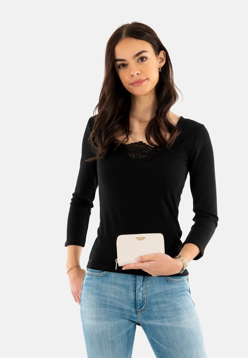 Camisa negra de manga larga con cuello de encaje, combinada con jeans de color azul claro. Sosteniendo una cartera cremosa con cremallera y patrón acolchado.